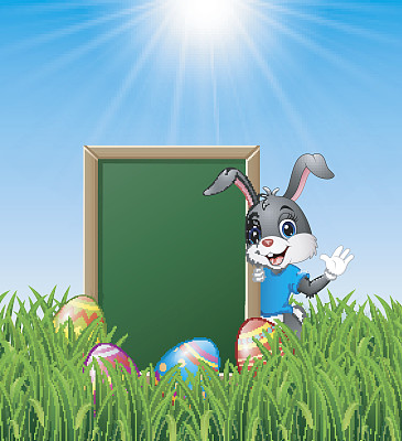 卡通兔子挥舞着复活节彩蛋和绿色黑板在草地上素材图片