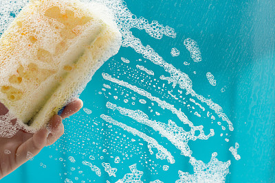 女性用黄色海绵和洗涤剂清洗窗户。玻璃上有肥皂污渍。家庭清洁的概念。图片下载