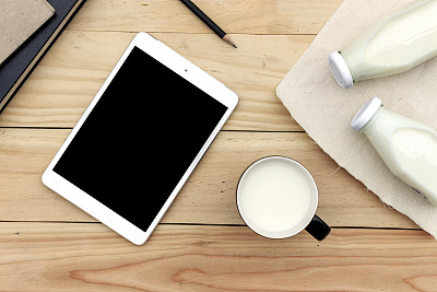 白色木桌上放着一杯牛奶、一台平板电脑和一本书，背景是软焦点。在光。乡村风格。图片下载