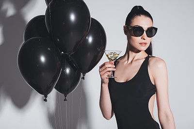 漂亮的年轻女人戴着太阳镜和泳衣喝马提尼，而站在工作室与黑色气球图片下载