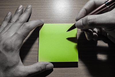 放在桌上的黄色记事本被手拿着并在上面写字。除了纸张的颜色是黄色外，色调是苍白的。这张便条提醒您的活动和活动不要忘记图片下载