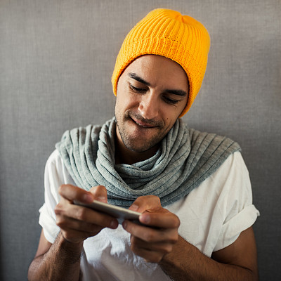 有吸引力的年轻人玩游戏的设备，电话。玩家戴着橙色帽子和灰色围巾，背景是灰色。被游戏迷住了。图片下载