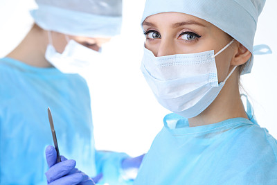 近距离的外科医生妇女看着摄像机，而同事在手术室的背景，孤立图片下载