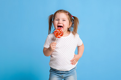 美丽的小女孩拿着巨大的螺旋形棒棒糖，在蓝色的背景上快乐地微笑着图片下载