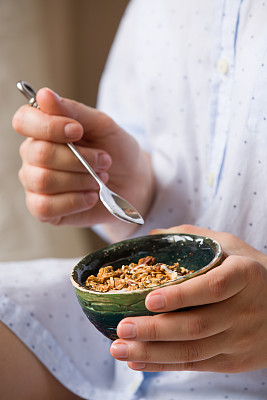 拿着麦片碗的年轻女子。女孩早餐吃麦片和坚果，南瓜子，燕麦和酸奶在碗里。拿着自制麦片的女孩。早上吃健康的零食或早餐。图片下载