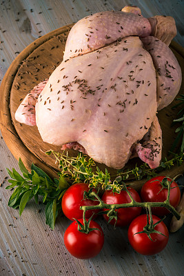 整只生鸡肉的垂直照片。肉的皮上有一种香料，如胡椒或孜然。很少有香草像百里香和迷迭香那样环绕着。鸡肉和蔬菜放在圆形木板上。图片下载