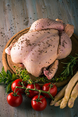 整只生鸡肉的垂直照片。肉的皮上有一种香料，如胡椒或孜然。很少有香草像百里香和迷迭香那样环绕着。鸡肉和蔬菜放在圆形木板上。图片下载
