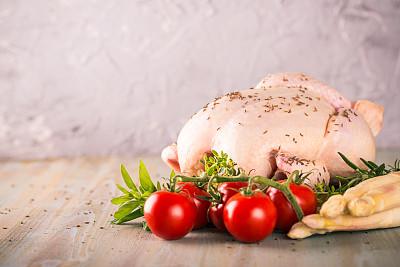 整只生鸡肉的水平照片。肉的皮上有一种香料，如胡椒或孜然。很少有香草像百里香和迷迭香那样环绕着。鸡肉和蔬菜放在圆形木板上。图片下载