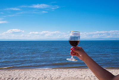 一个做了漂亮美甲的女人手里拿着一杯红酒。以阳光明媚的海滩为背景。蓝色的大海和蓝色的天空。图片下载