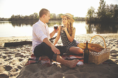非常爱你。这是一对美丽的年轻夫妇手拿酒杯，坐在海滩上浪漫野餐时互相看着对方的照片。图片下载