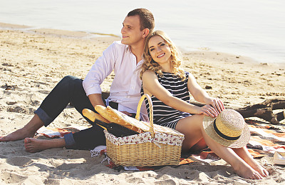爱的夫妻。水平的照片美丽的年轻女人看着相机和微笑，而坐在海滩上的男朋友和野餐。图片下载