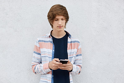 年轻的潮男，留着时髦的发型，专注地看着他的智能手机，使用免费互联网连接在线下载图片。用手机上网的男生图片下载