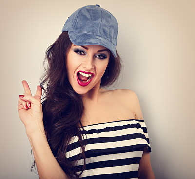 疯狂的年轻女模特在蓝色的帽子显示岩石姿态与张开的嘴在蓝色的背景与空白的复制空间。明亮的妆容和红色的口红。特写镜头的肖像图片下载