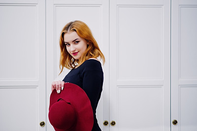 肖像时尚红发女孩的红帽子和黑色连衣裙与明亮的化妆摆近白色古董木制橱柜。照片色调风格的Instagram滤镜。图片下载