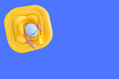 婴儿漂浮在黄色的充气筏上。小孩用铲子在黄色橡皮环上一个蓝色的背景与拷贝空间。从上面看。图片下载