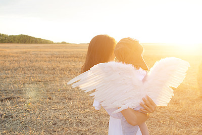 母亲和儿子玩得很开心。男孩戴着飞行员帽，带着天使翅膀坐在他妈妈的手上看日落。图片下载