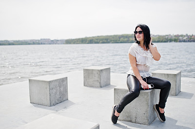 照片中的黑发女孩戴着耳机听着手机里的音乐，穿着女人的皮裤和白衬衫，戴着太阳镜，在湖边的沙滩上倚着石方。图片下载