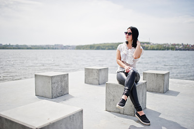 照片中的黑发女孩戴着耳机听着手机里的音乐，穿着女人的皮裤和白衬衫，戴着太阳镜，在湖边的沙滩上倚着石方。图片下载
