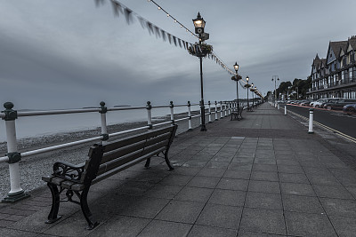 这是一幅英国海滨柔和的画面，长椅俯瞰着海滩和大海，灯柱之间飘扬着彩旗。图片下载