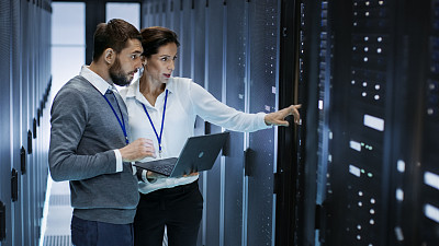 男性IT专家拿着笔记本电脑，与女性服务器技术人员讨论工作。他们站在数据中心，机架服务器柜是开放的。图片下载