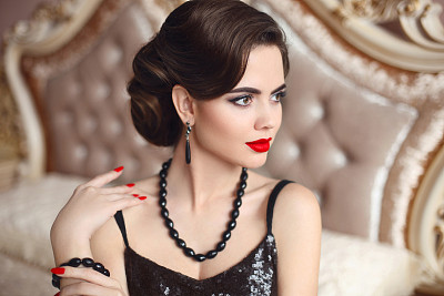 美丽的黑发，优雅的女人肖像。复古女士红唇妆，波浪发型和时尚的黑宝石珠宝摆姿势在现代卧室室内的床上。奢侈的生活。图片下载