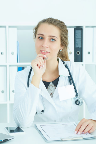一个自信的年轻女医生坐在桌旁看着镜头的肖像。医生在等病人检查。医生接待的概念图片下载