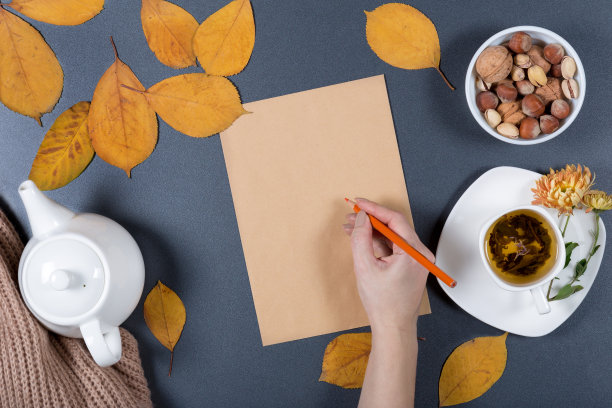 秋天的工作桌。黄色和橙色的叶子，白色的一杯凉茶，茶壶，牛皮纸和坚果。健康的零食。女人的手拿着铅笔，写着纸条。图片下载