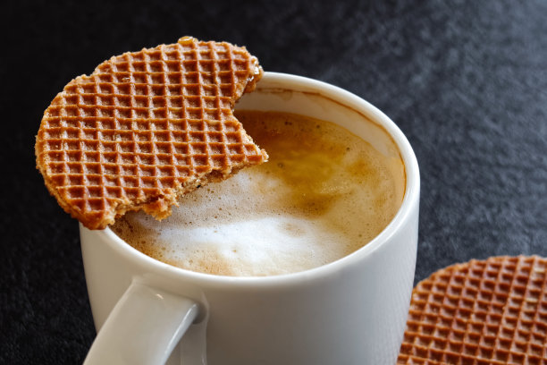 白色杯子上有半喝半喝的奶泡咖啡和半吃半吃的华夫饼干。黑色背景。图片下载