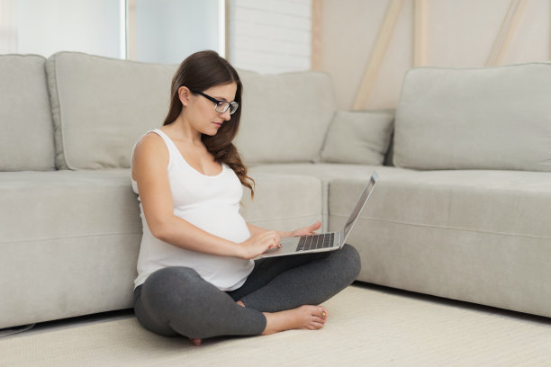 一名孕妇坐在家里明亮的地板上。她双腿交叉坐在地板上，在笔记本电脑上工作图片下载