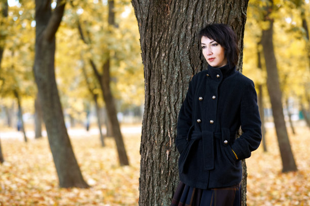 美丽的女孩站在秋城公园的树旁，黄叶为背景，秋季，穿着黑色的外套图片下载