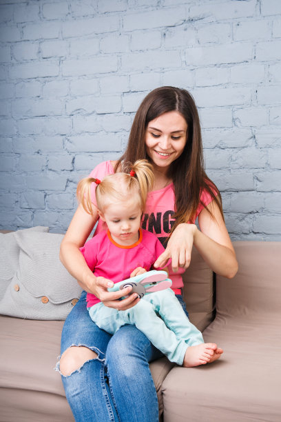 美丽的年轻母亲用智能手机玩一个两岁的女儿坐在沙发上在房间里穿着明亮的颜色，彩色的衣服图片下载