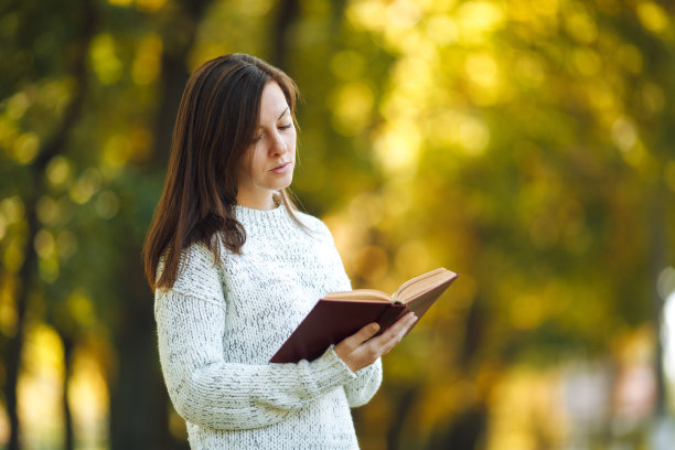 一个美丽的棕色头发的女人在一个温暖的日子里站在秋天的城市公园里，穿着白色的毛衣，拿着一本红色的书。秋天金色的叶子。阅读的概念。图片下载