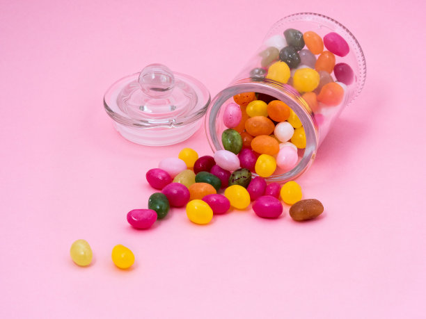 从带盖的玻璃罐中，不同颜色的果冻糖果分散在粉红色的背景上。染料和防腐剂。图片下载