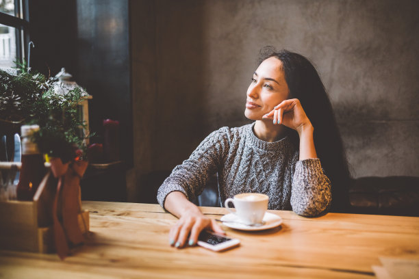 在一家装饰着圣诞装饰的咖啡馆里，美丽的年轻女孩拿着一个白色的杯子喝咖啡，旁边放着她的手机。穿着一件灰色针织羊毛衫图片下载