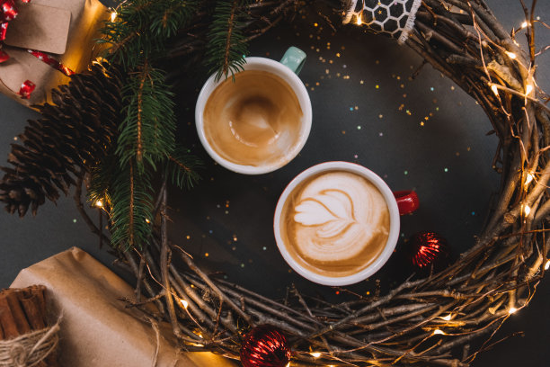 新鲜美味的早晨，两杯卡布奇诺(其中一杯已经空了)，手工制作的木制圣诞花环，上面有萤火虫、云杉树枝和灰色背景上的金色闪光图片下载