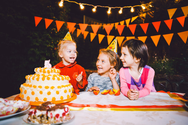 孩子的生日聚会。三个欢快的小女孩坐在桌前用手抹着脸吃蛋糕。用明亮的灯泡装饰庭院，营造欢乐喜庆的气氛图片下载