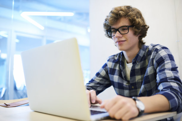 IT公司的年轻男性实习生戴着时髦的眼镜，通过笔记本电脑和免费的5G无线网络练习编程，以提高技能，成为成功的软件开发人员图片下载