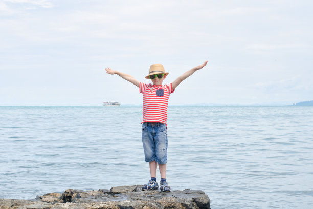 男孩穿着条纹t恤，戴着太阳镜，双手高举，背对着大海图片下载
