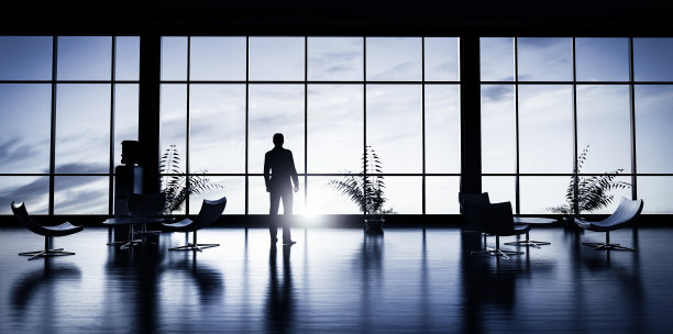 一个商人站在办公室里看着窗外日落的天空。图片下载