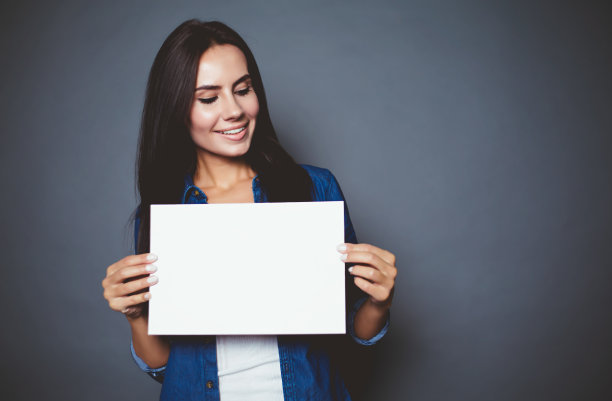 美丽的现代微笑的女人在牛仔裤衬衫与空白纸广告在一个灰色的背景孤立。素材图片