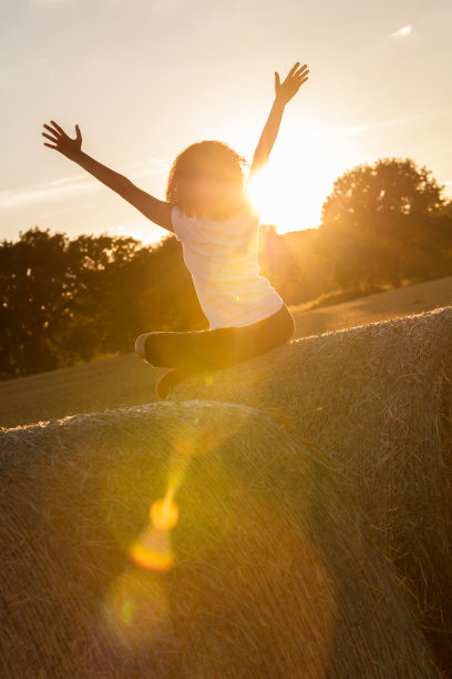 混血非洲裔美国女孩年轻女性坐在干草捆上双臂举起庆祝日落或日出金色的傍晚或早晨的阳光图片下载
