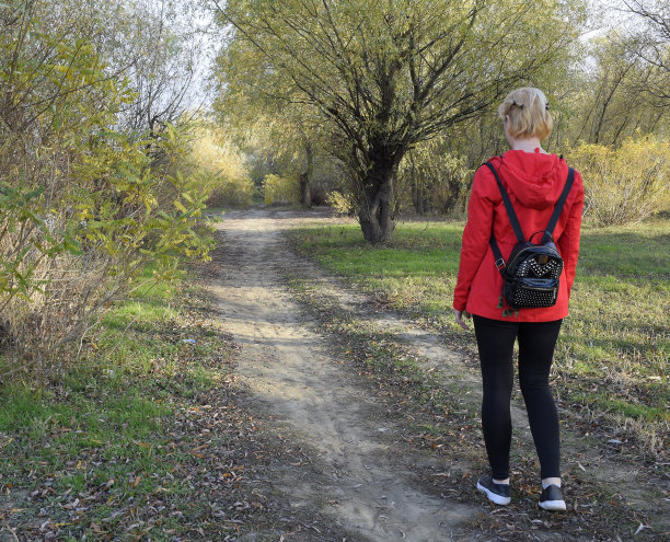 女孩穿着红色的夹克和一个背包沿着路去森林。路的开始。图片下载