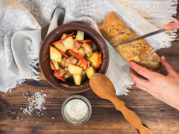 女人们的手在桌子的背景上切面包。乡村静物画，烤土豆配肉放在瓷碗里。面包和盐的背景木桌和粗糙的布，俯视图图片下载