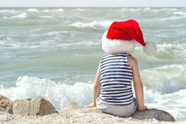 戴着圣诞帽坐在海滩上的孩子。后视图图片下载