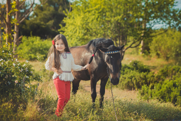 女孩有着深褐色的头发和棕色的眼睛，穿着时髦的穿着乡村衣服，白色的衬衫和红色的裤子在皮带上与黑色的小马驹摆姿势图片下载
