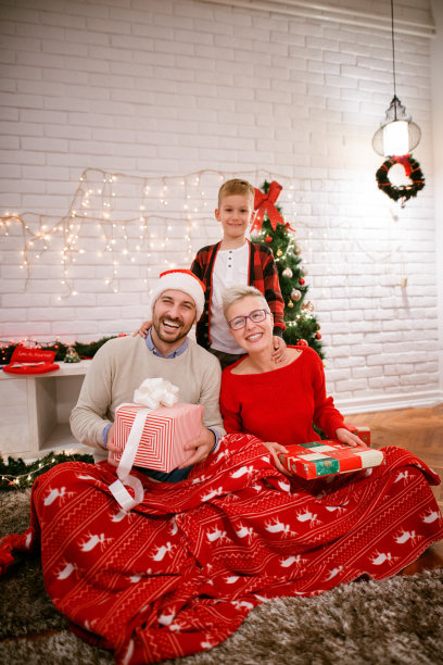 圣诞节的全家福照片在家里，而快乐满意的父母坐在地毯上的红毯，拿着礼物在可爱的小男孩面前看着相机图片下载