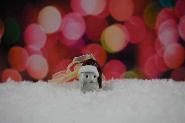 这是一幅可爱的圣诞照片，圣诞老人帽子上装饰着瓷制的白兔树，坐在雪地里，背景是温暖的红粉色仙女灯图片下载