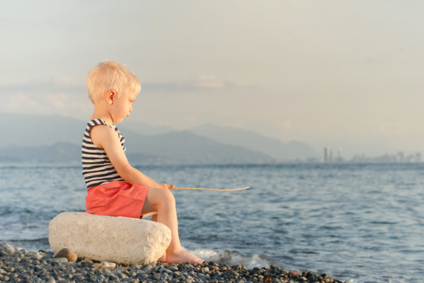 穿着条纹t恤的男孩拿着棍子坐在沙滩上。海、山、城市，远处的摩天大楼为背景图片下载