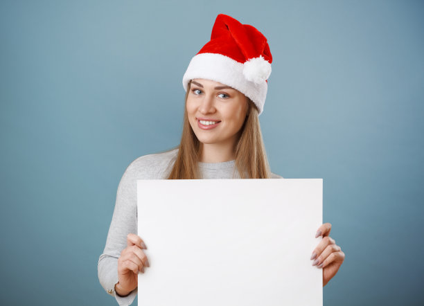 圣诞微笑的女孩拿着白色的大卡片。圣诞老人的帽子。图片下载
