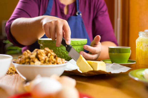 近距离的照片，年轻的女性坐在一个木制厨房桌子后面，专注于她的手拿着一把刀，切一片黄油。在桌子上烘焙的原料。图片下载